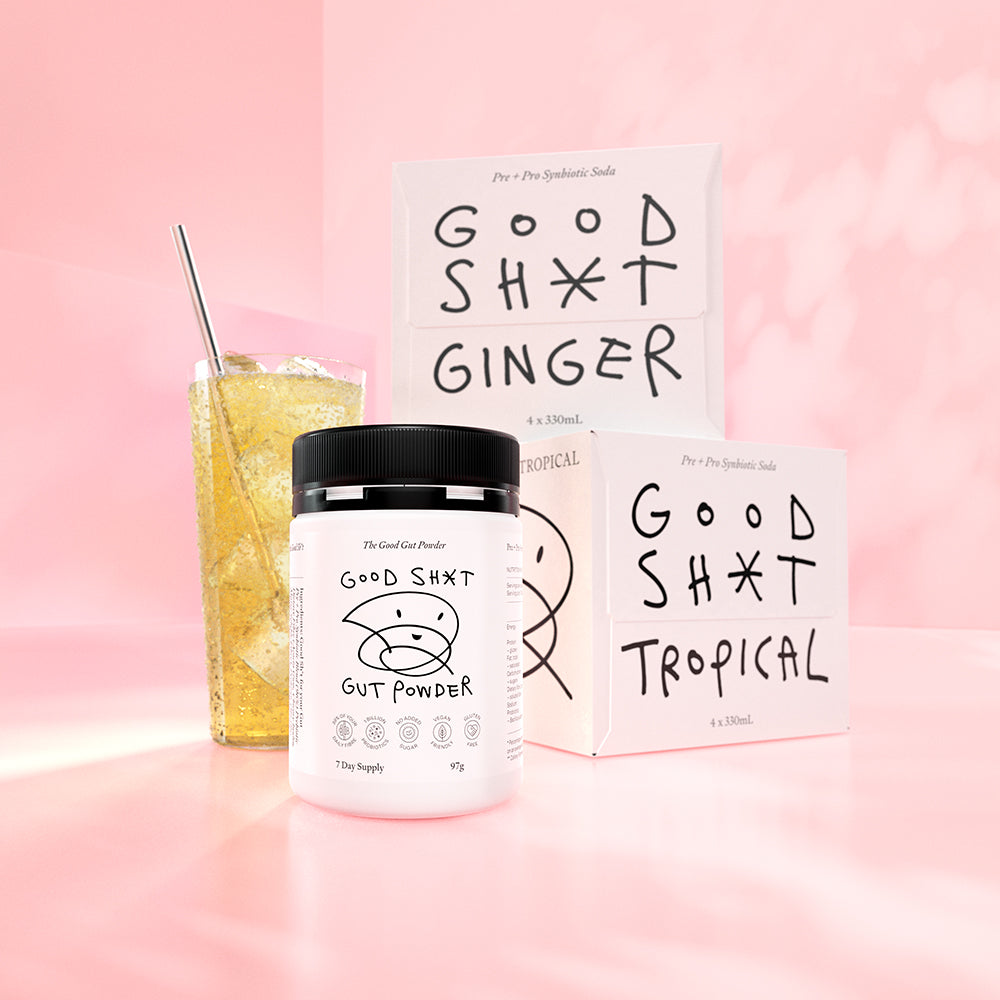 A stack of two 4 pack boxes of Good Shit pre + probiotic soda. One box each of ginger and tropical, plus a tub of Good Shit pre + probiotic Gut Powder, containing enough powder for 7 days, with a frosty glass of golden hued Good Shit ginger pre + probiotic soda with ice behind it